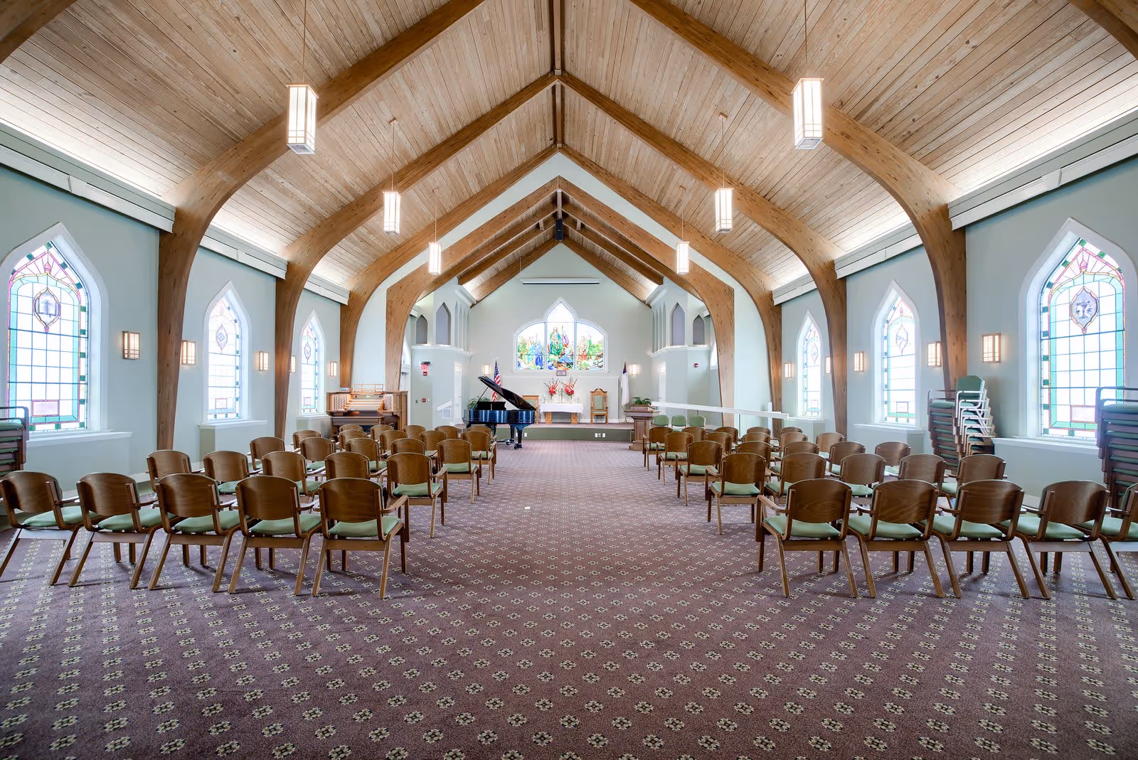Interior view of a chapel with wooden vaulted ceiling and stained glass windows on both sides. Rows of wooden chairs with green cushions are arranged facing an altar at the front, which features a piano, an organ, floral arrangements, and religious symbols.