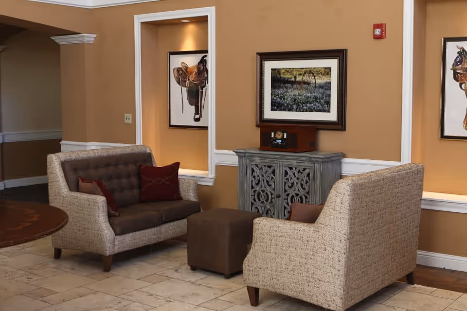 A cozy sitting area with two beige and brown upholstered loveseats facing each other, a small brown ottoman in between, a decorative cabinet with a vintage radio on top, and framed artwork on the tan walls.
