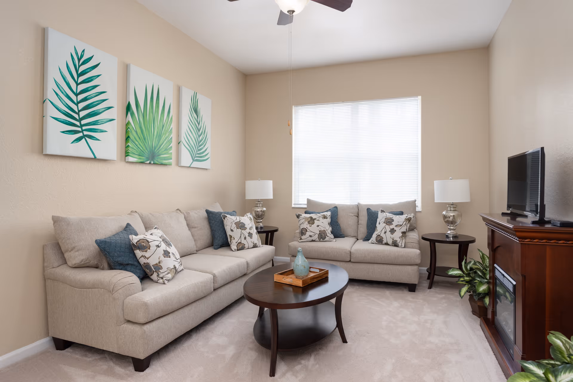 A cozy living room with two beige sofas adorned with floral and blue cushions, a wooden oval coffee table with a decorative tray and vase, two side tables each with a lamp, a flat-screen TV on a wooden stand with a built-in fireplace, and three framed botanical prints on the wall. The room has beige walls, a large window with blinds, and a ceiling fan.