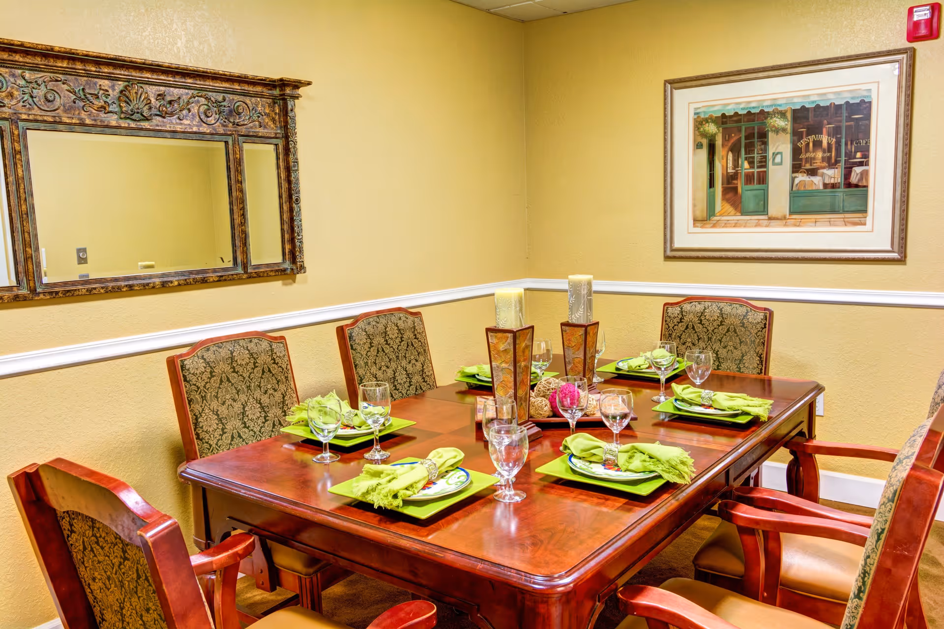 A formal dining room with a wooden table set for six people. Each place setting includes a green napkin, a plate, and a glass. The room has beige walls with white trim, a large decorative mirror on one wall, and a framed painting of a storefront on another wall. The chairs around the table have patterned upholstery and wooden frames.
