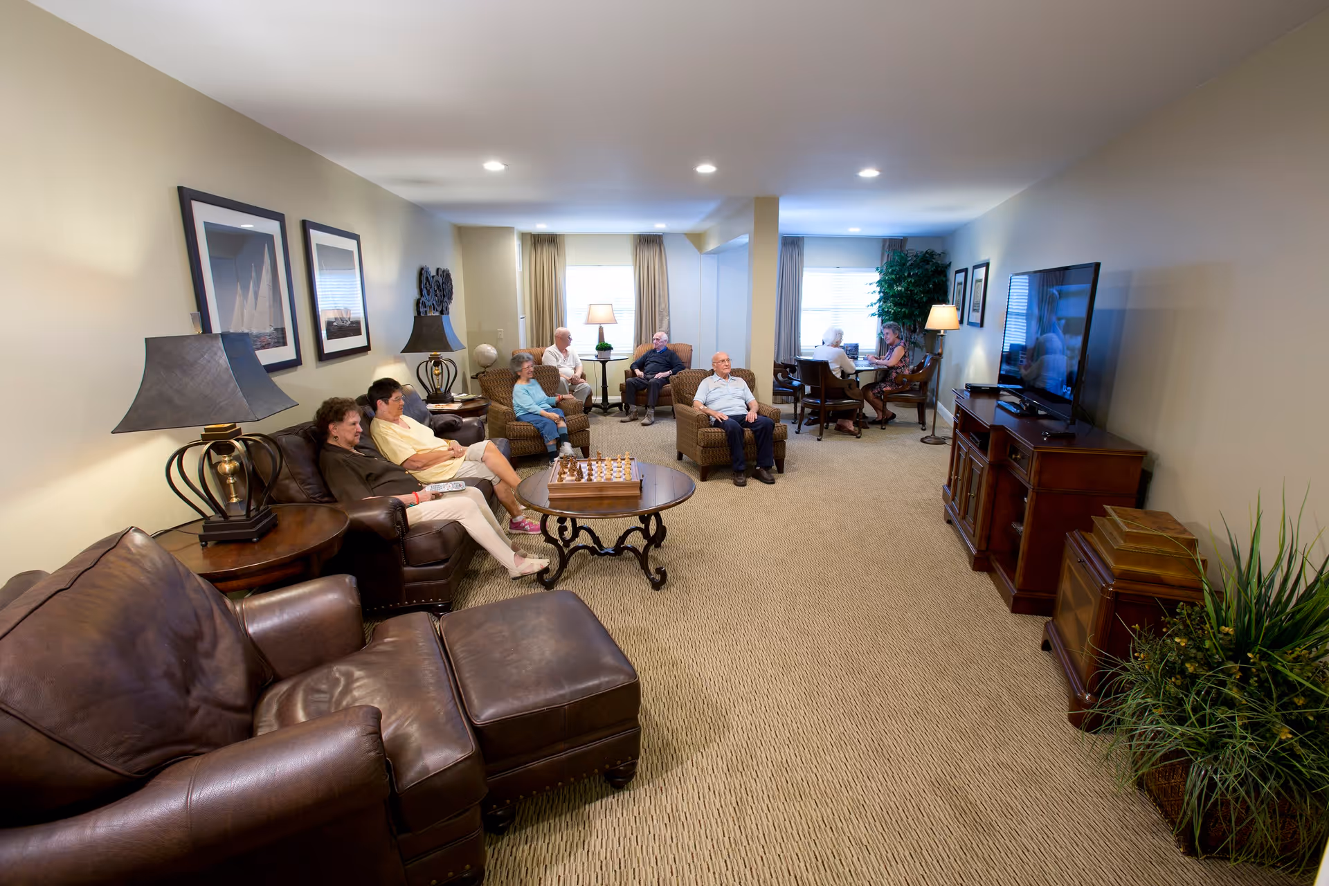 A spacious living room in Armbrook Village with several elderly people sitting on leather and fabric chairs arranged around a coffee table with a chess set. The room has beige walls, carpeted floors, framed pictures on the walls, table lamps, a large flat-screen TV on a wooden cabinet, and large windows with curtains allowing natural light.