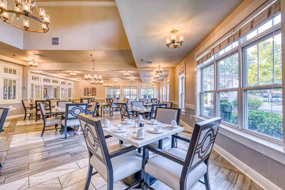 Bright and spacious dining room in Chesapeake Place Senior Living with multiple tables set with white dishes, cups, and utensils. The room features large windows allowing natural light to fill the space, elegant chandeliers hanging from the ceiling, and comfortable chairs with decorative backs arranged around the tables.