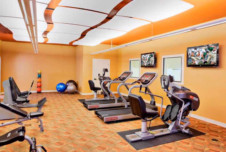 Brightly lit fitness room with treadmills, recumbent bikes, exercise balls and wall-mounted TVs.