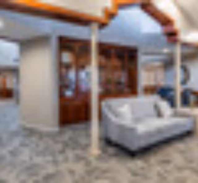 A cozy living room area featuring a light gray sofa with white pillows, wooden beams on the ceiling, and a built-in wooden cabinet with glass doors. The floor is carpeted with a patterned design, and the space is softly lit with natural and artificial light.