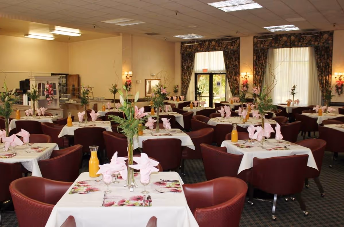 A dining room with multiple tables covered in white tablecloths, each set with pink folded napkins, floral placemats, and a vase with flowers and greenery. There are maroon upholstered chairs around each table. The room has large windows with floral curtains and wall sconces with lights.