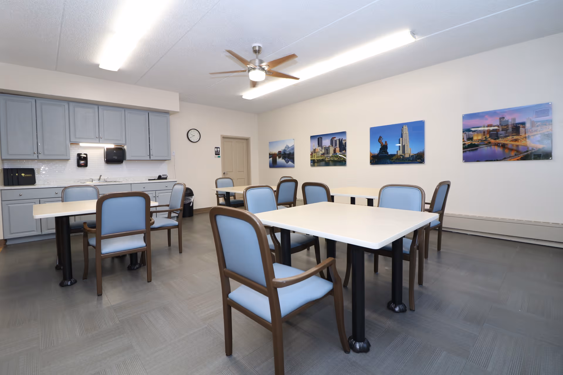 A dining room with several tables and chairs arranged neatly. The chairs have light blue cushions and wooden frames. On the left side, there is a kitchenette area with gray cabinets, a sink, and a paper towel dispenser. The walls are decorated with framed cityscape photographs. The room is well-lit with ceiling lights and a ceiling fan.