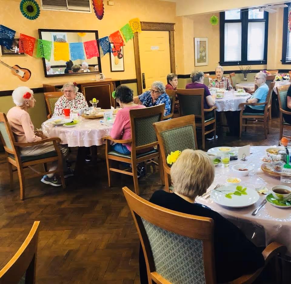 A group of elderly people sitting around tables in a decorated dining room, engaging in conversation. The room has yellow walls, wooden chairs, and colorful paper decorations hanging on the wall. Plates, cups, and food are on the tables.