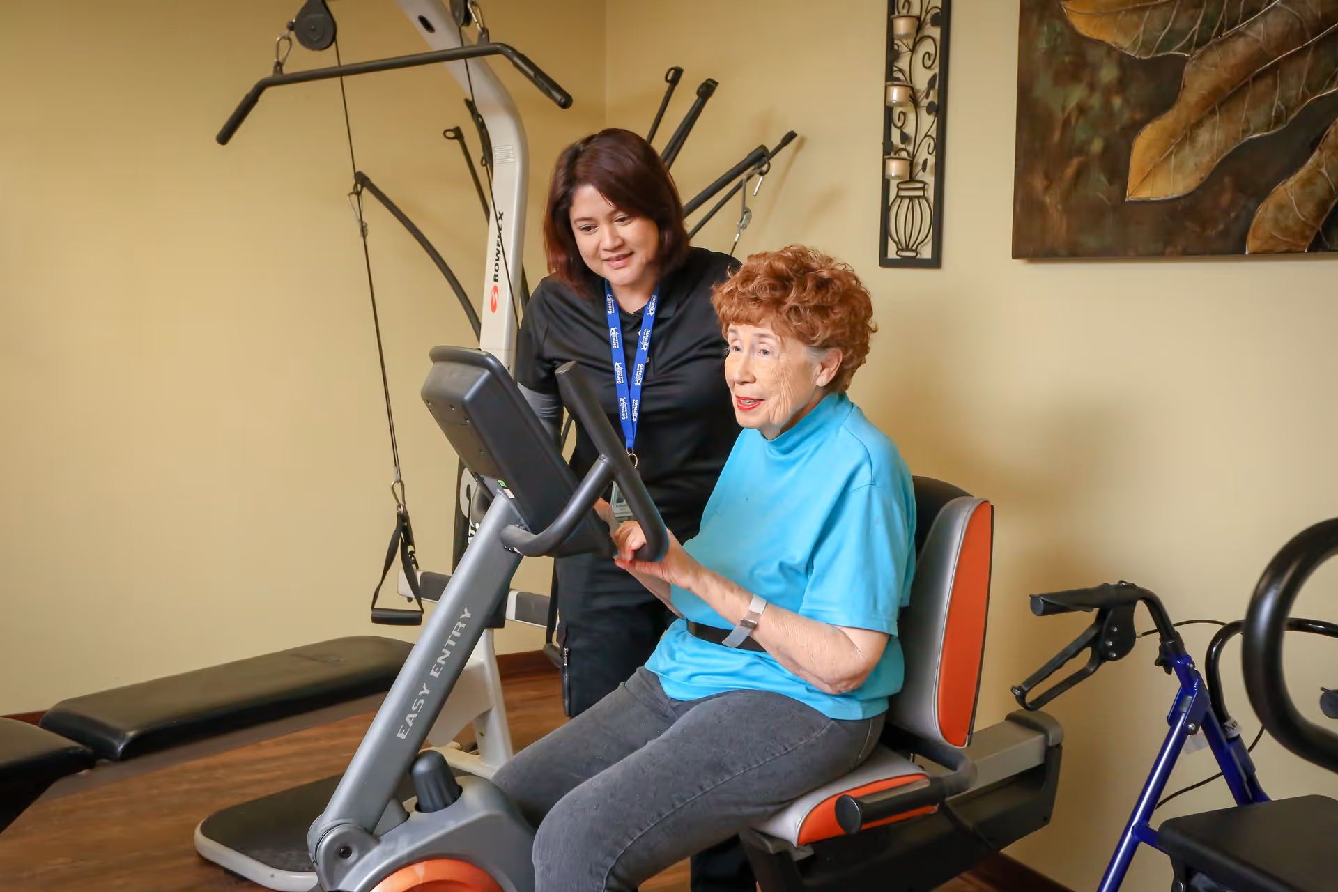 An elderly woman in a blue shirt is seated on a recumbent exercise bike labeled 'EASY ENTRY' while a female caregiver or trainer stands beside her, offering support and encouragement in a fitness or rehabilitation room with exercise equipment and a walker nearby.