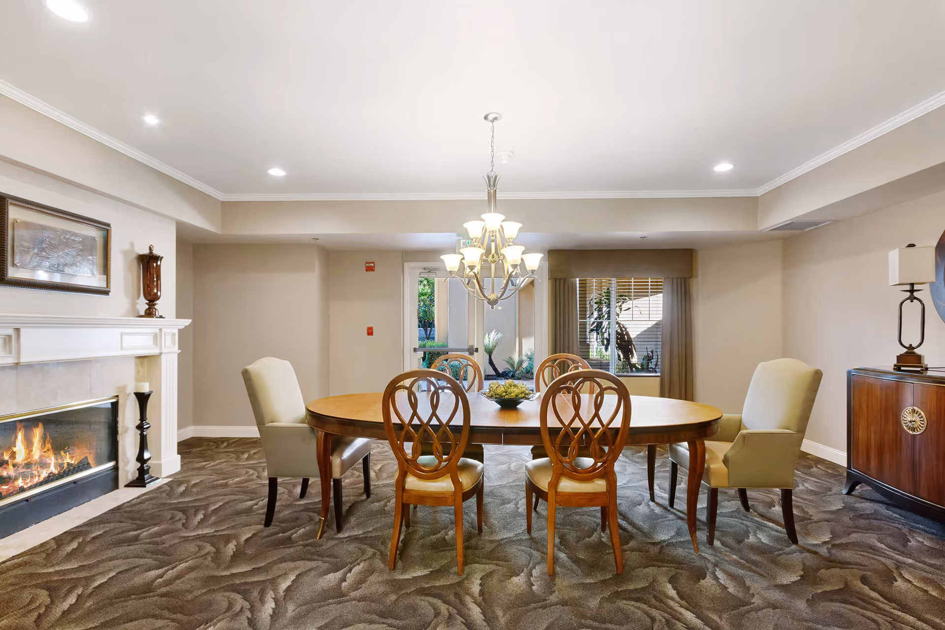 A cozy dining room with a wooden oval table surrounded by six chairs, two upholstered armchairs and four wooden chairs with decorative backs. A lit fireplace with a white mantel is on the left wall, and a chandelier with multiple lights hangs above the table. There is a window with blinds and a door leading outside at the back of the room. The room has beige walls and patterned carpet flooring.