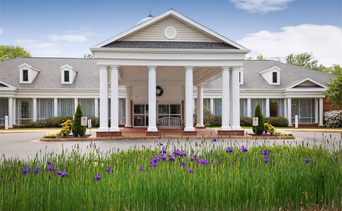 Front entrance of a white-columned building with a covered porte-cochère and purple flowers in the foreground.