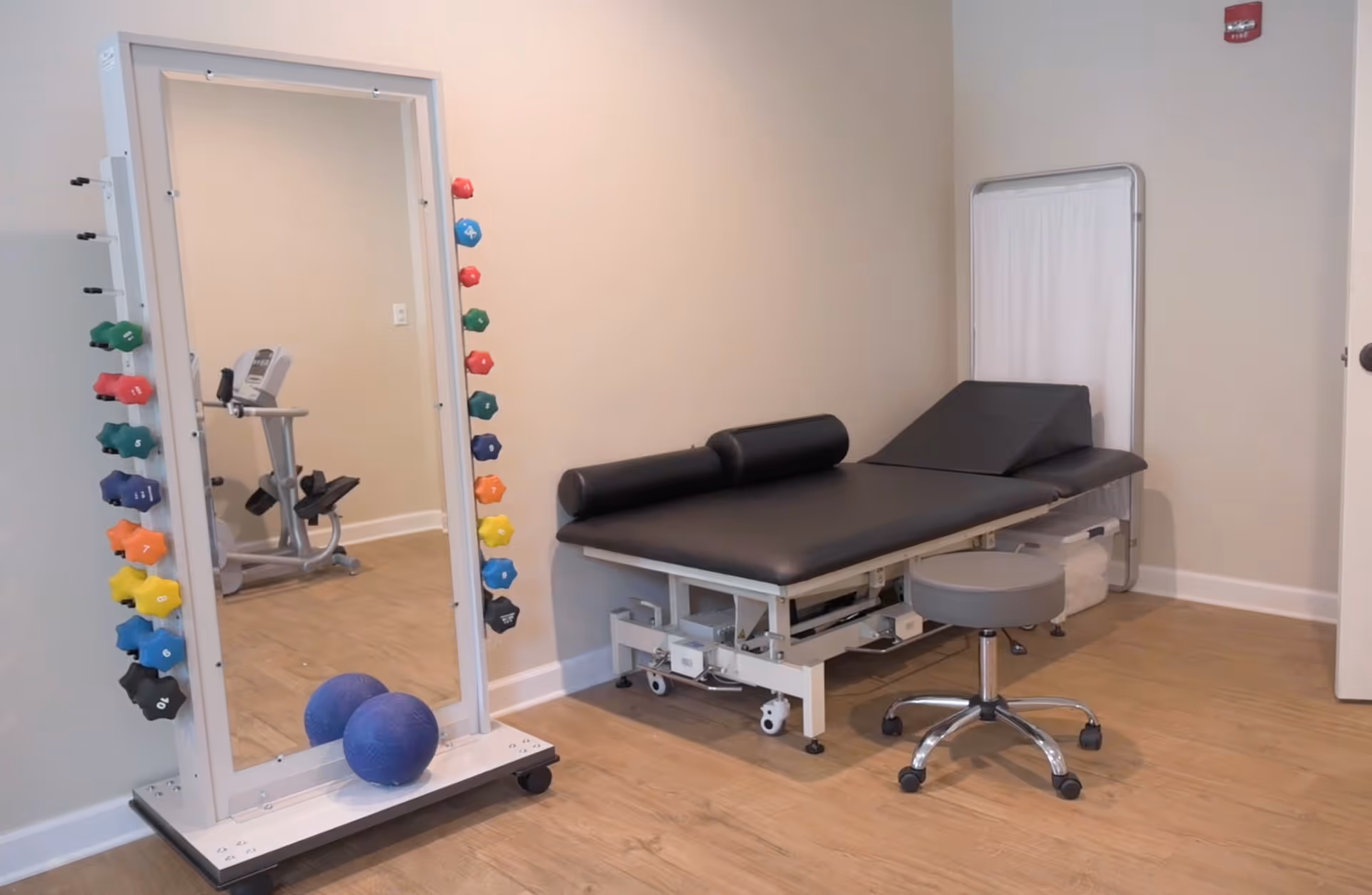 A therapy or examination room with a black padded adjustable treatment table, a gray rolling stool, a large mirror with colorful dumbbells attached to its sides, and an exercise bike reflected in the mirror. The room has light-colored walls and wood flooring.
