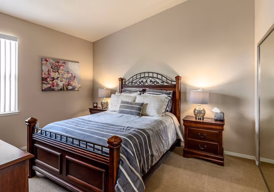 A neatly made bed with a dark wooden frame and metal decorative headboard in a bedroom. The bed has striped bedding in shades of gray and white, with multiple pillows. On each side of the bed are matching wooden nightstands with lamps, a clock, and a tissue box. A floral painting hangs on the beige wall to the left, and a window with vertical blinds is partially visible. A mirrored closet door is on the right side of the room.