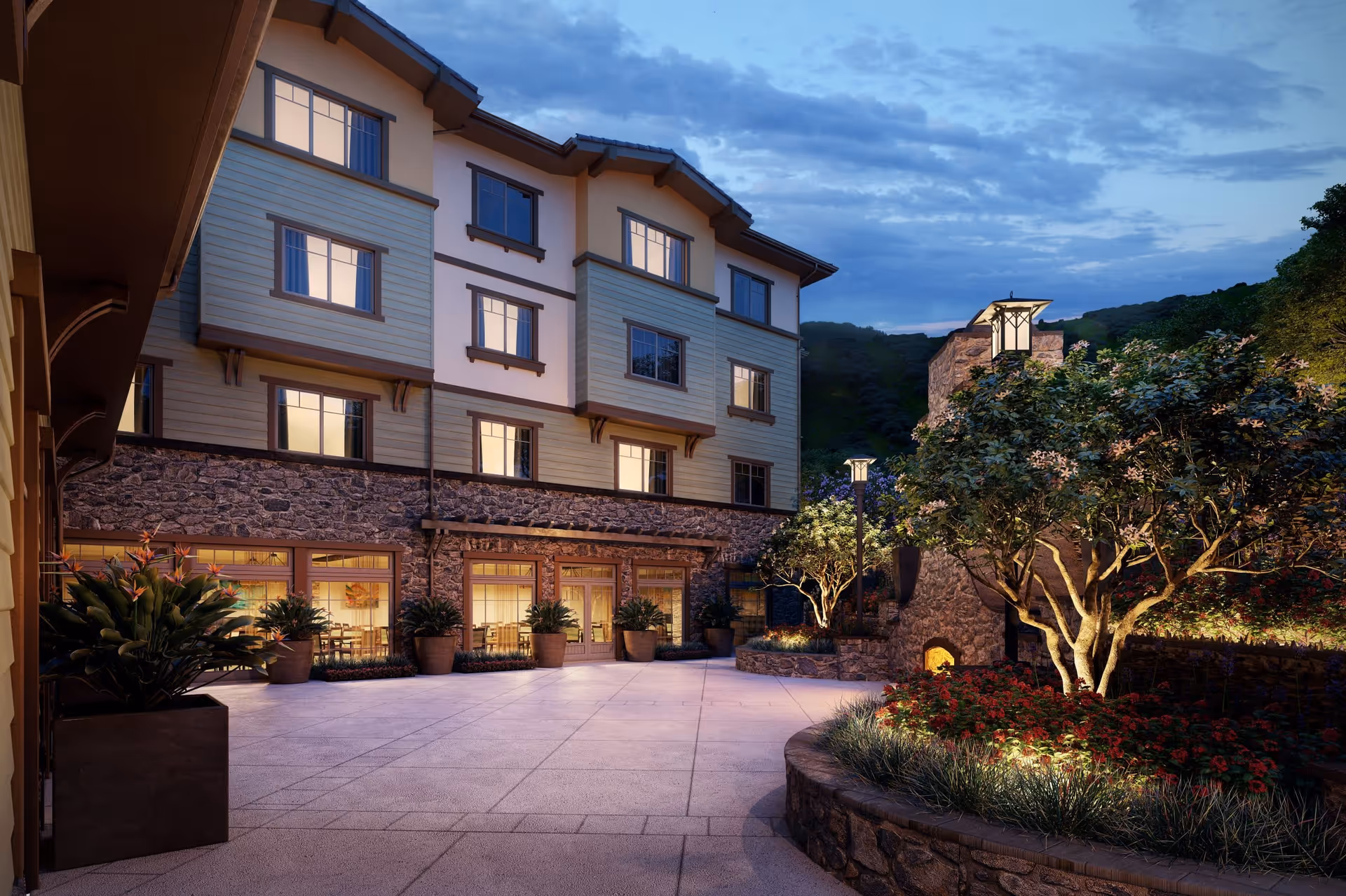 Evening view of an outdoor courtyard area at Sage Mountain facility featuring stone and wood exterior walls, large windows with warm interior lighting, potted plants, a stone fireplace, and landscaped garden beds with trees and flowers under a partly cloudy sky.