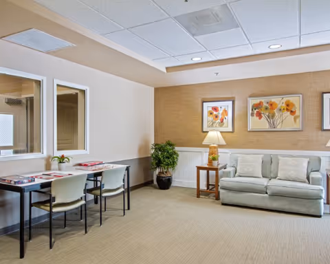 A cozy common area with a light green sofa, a wooden side table with a lamp, and two framed floral paintings on the wall. There is a small table with two chairs along the opposite wall, decorated with a small plant and some magazines. A potted plant is placed in the corner, and the room has beige carpeting and a drop ceiling with recessed lighting.