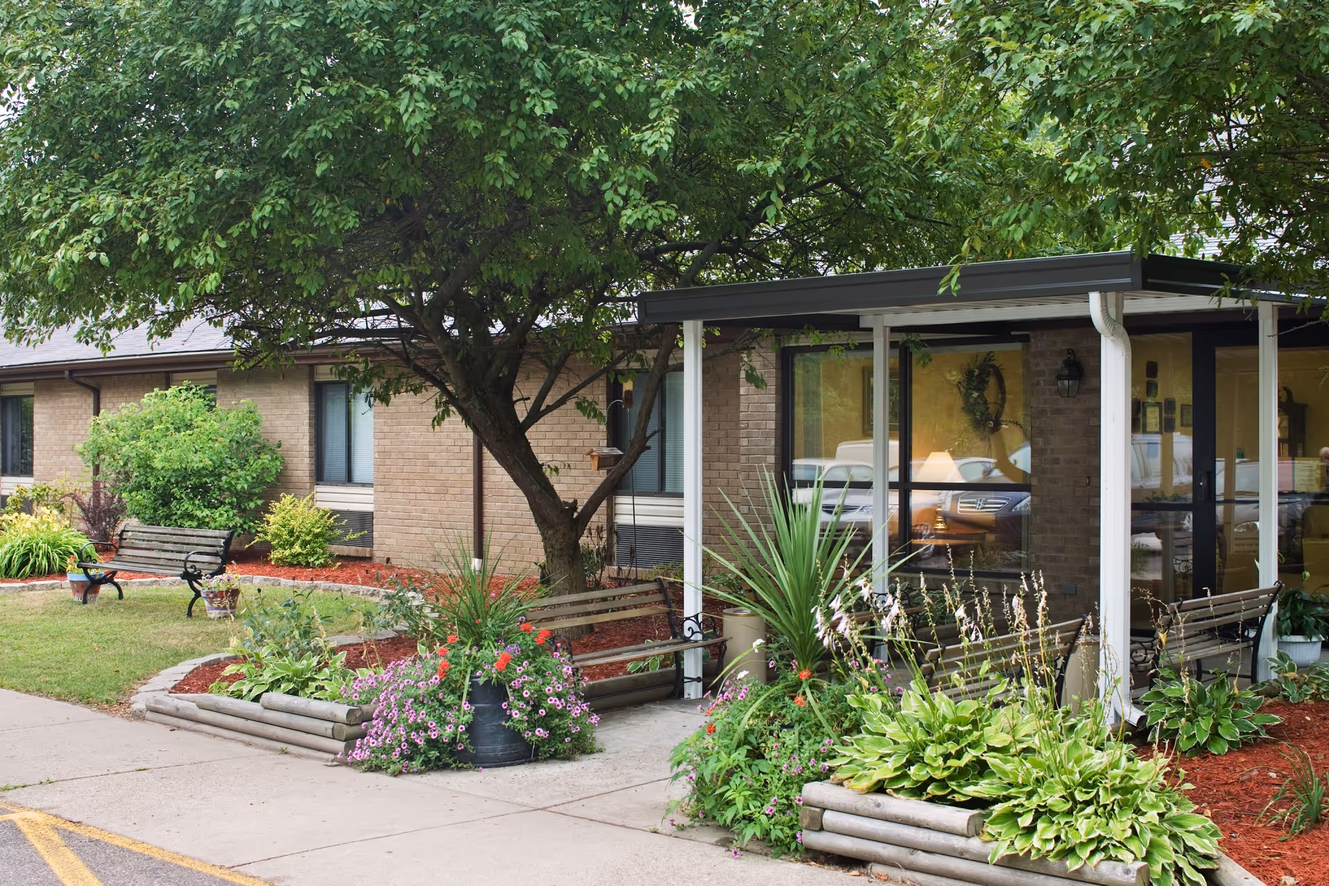 Outdoor garden area at Teays Valley Center featuring a large tree, flower beds with various plants and flowers, wooden benches, and a covered entrance to a brick building with large windows.