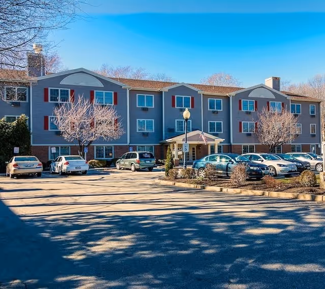 Exterior view of a three-story senior living facility building with gray siding and red shutters. Several cars are parked in the parking lot in front of the building, and there are leafless trees and small shrubs around the entrance area under a clear blue sky.
