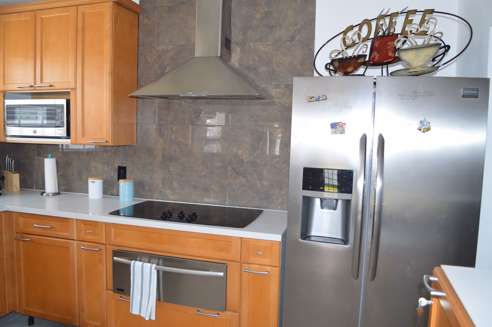Modern kitchen with wooden cabinets, a stainless steel refrigerator with magnets, a built-in microwave, an electric stovetop with a stainless steel range hood, and a white countertop with kitchen utensils and containers.
