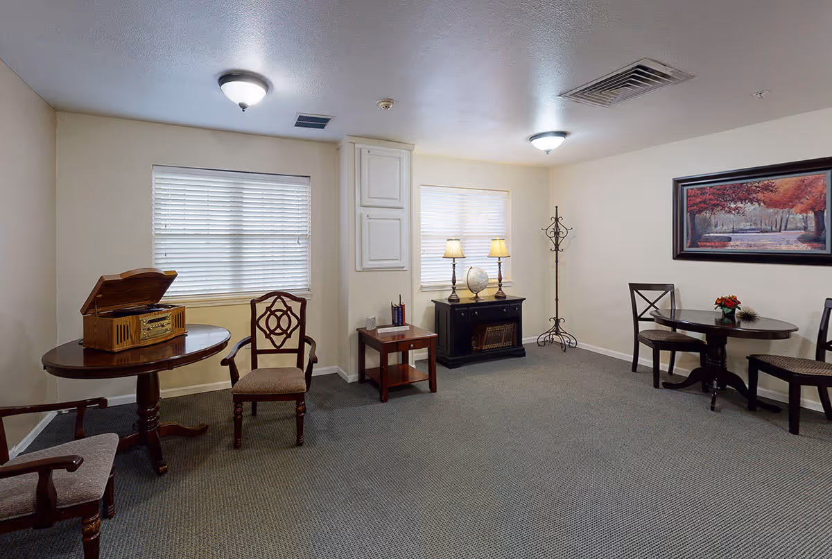 A cozy common area with beige walls and carpeted floor featuring two round wooden tables with chairs around them. One table has a vintage-style record player, and the other has a small flower arrangement. There are two windows with blinds, a small side table with books, a black cabinet with two lamps and a globe, a coat rack, and a framed painting of a tree-lined path with autumn foliage on the wall.