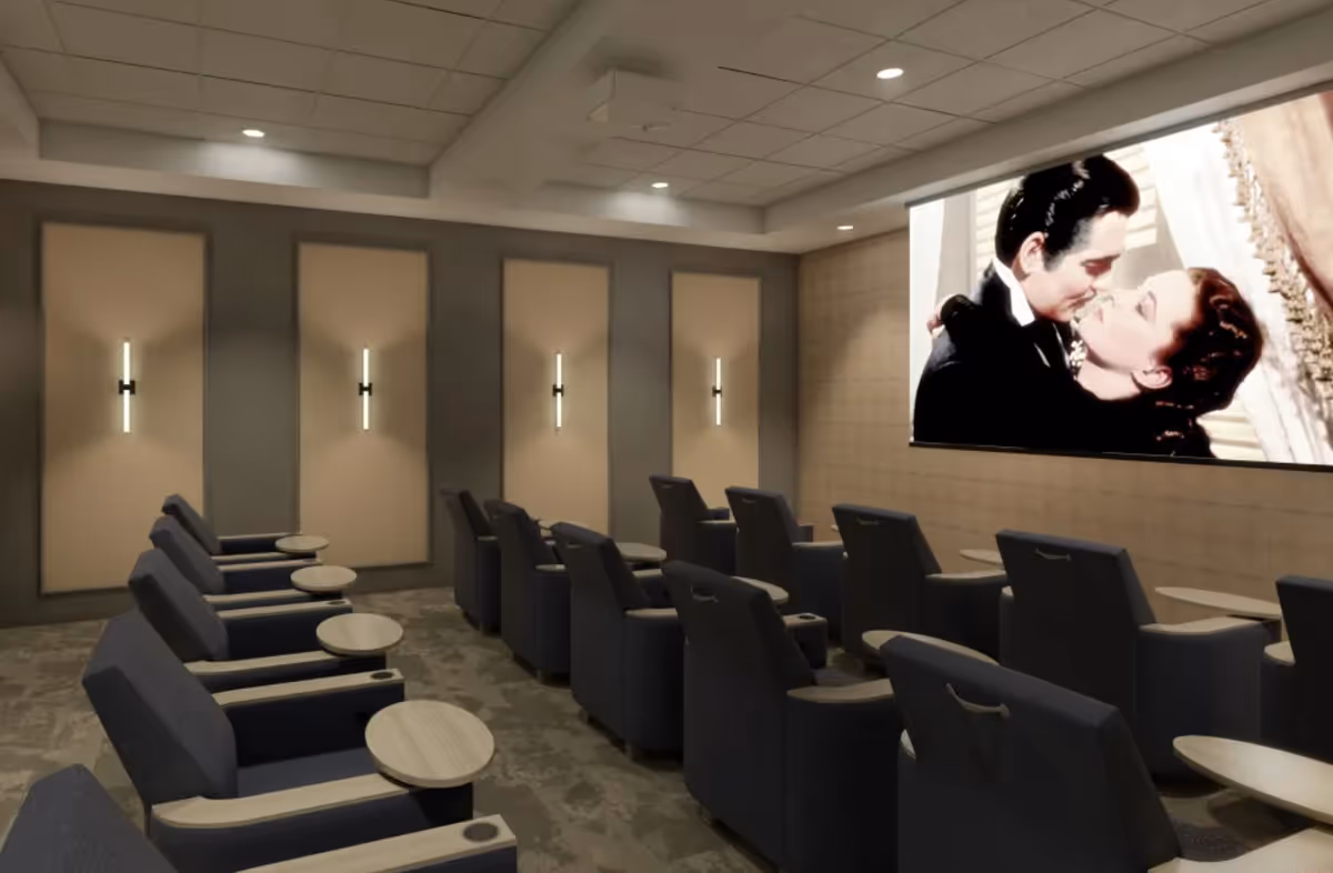 A small movie theater room with two rows of dark gray reclining chairs, each equipped with a small wooden side table. The room has beige walls with vertical light fixtures and a large screen showing a classic black-and-white movie scene of a man and woman embracing.