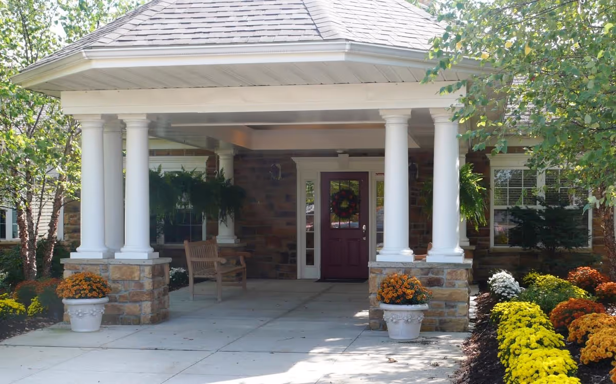 Covered front entrance with white columns, a maroon door, bench and potted flowers surrounded by landscaped beds.