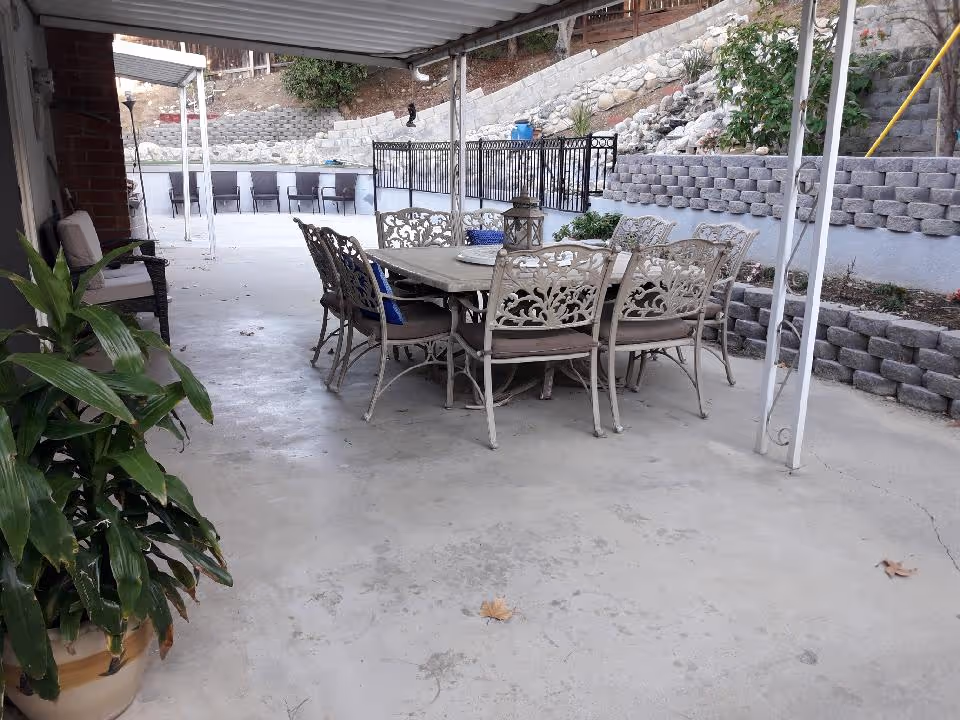 Covered outdoor patio with a metal dining table and chairs, a potted plant, and terraced stone retaining walls.