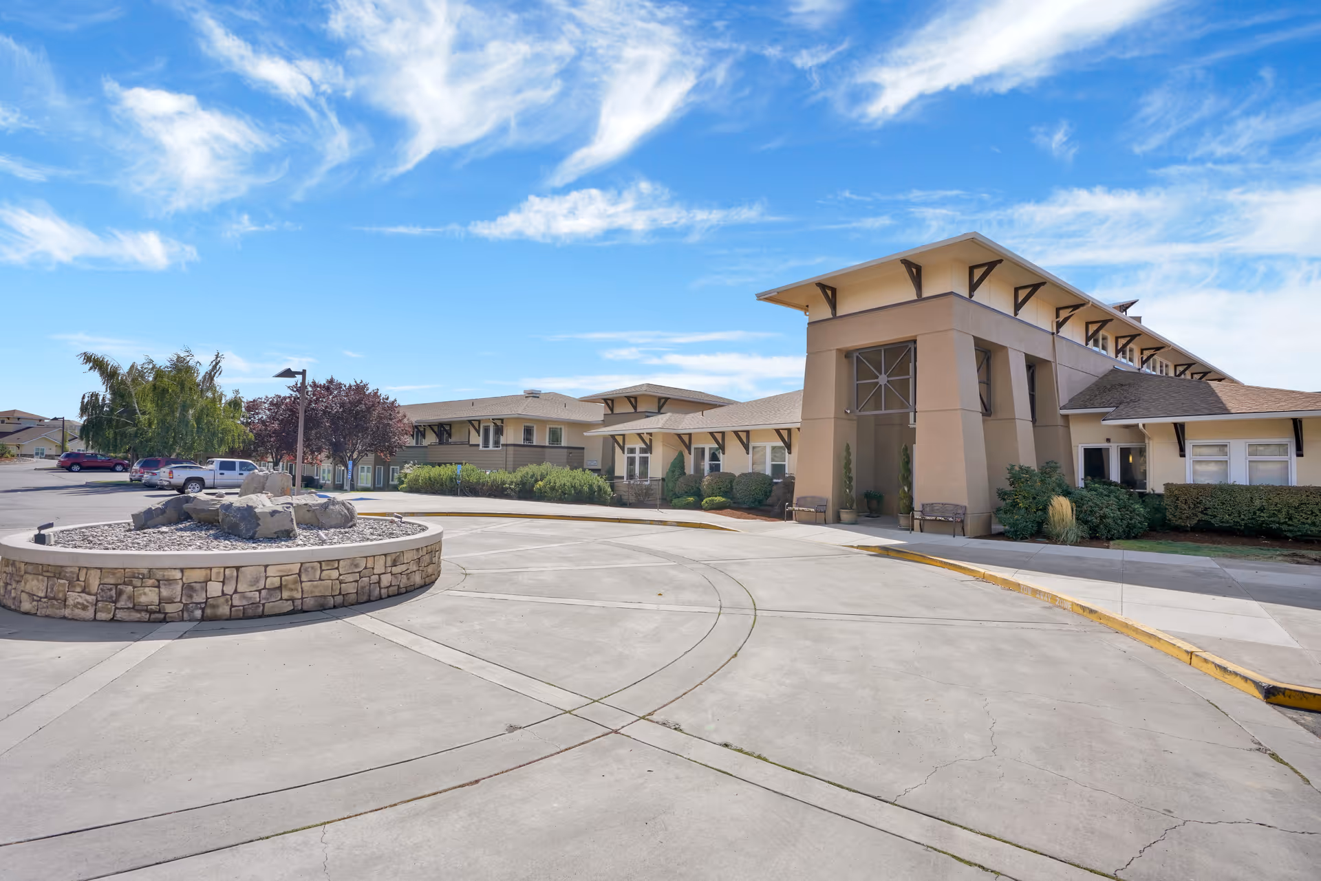 Exterior view of Crystal Terrace of Klamath Falls senior living facility showing a large circular driveway with a stone planter in the center, surrounded by a beige building with multiple windows and a clear blue sky with some clouds.