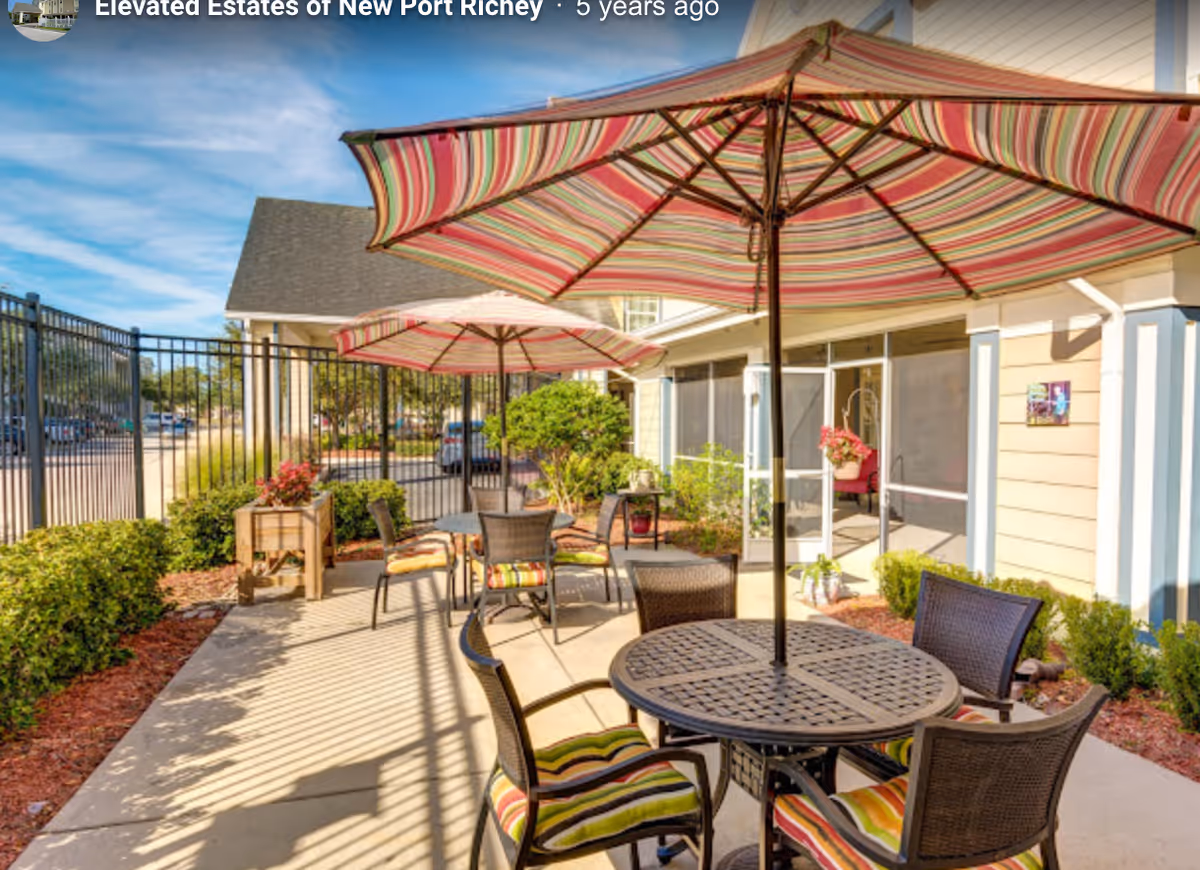 Outdoor patio with round tables, striped umbrellas and chairs next to a senior living building.