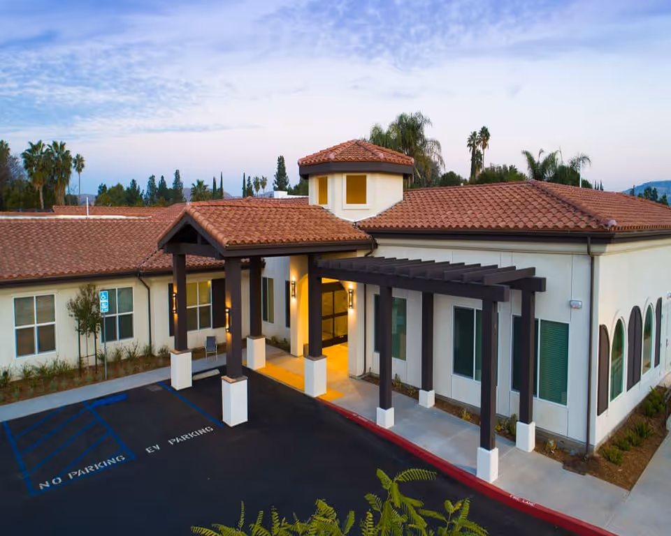Exterior view of The Preserve at Woodland Hills Assisted Living & Memory Care building entrance with a covered walkway, tiled roof, and surrounding greenery under a partly cloudy sky.
