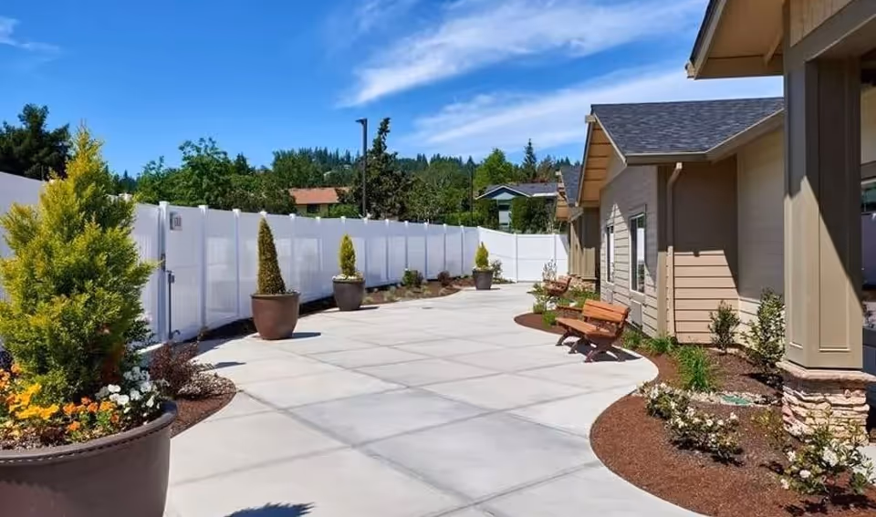 Outdoor paved walkway area at Sunnyside Meadows Memory Care with benches, potted plants, and landscaped garden beds along the side of a building under a blue sky.