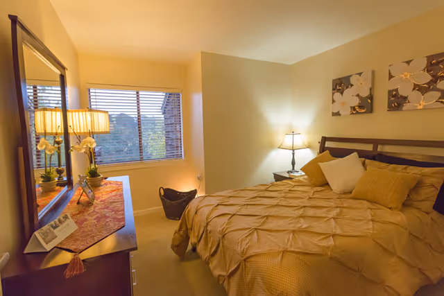 A cozy bedroom with a large bed covered in a beige textured comforter and multiple pillows. There is a wooden nightstand with a lamp on the right side of the bed. On the left side of the room, a wooden dresser with a mirror above it holds a table lamp, a small plant, and some decorative items. A window with horizontal blinds lets in natural light, and two floral paintings hang on the wall above the bed.