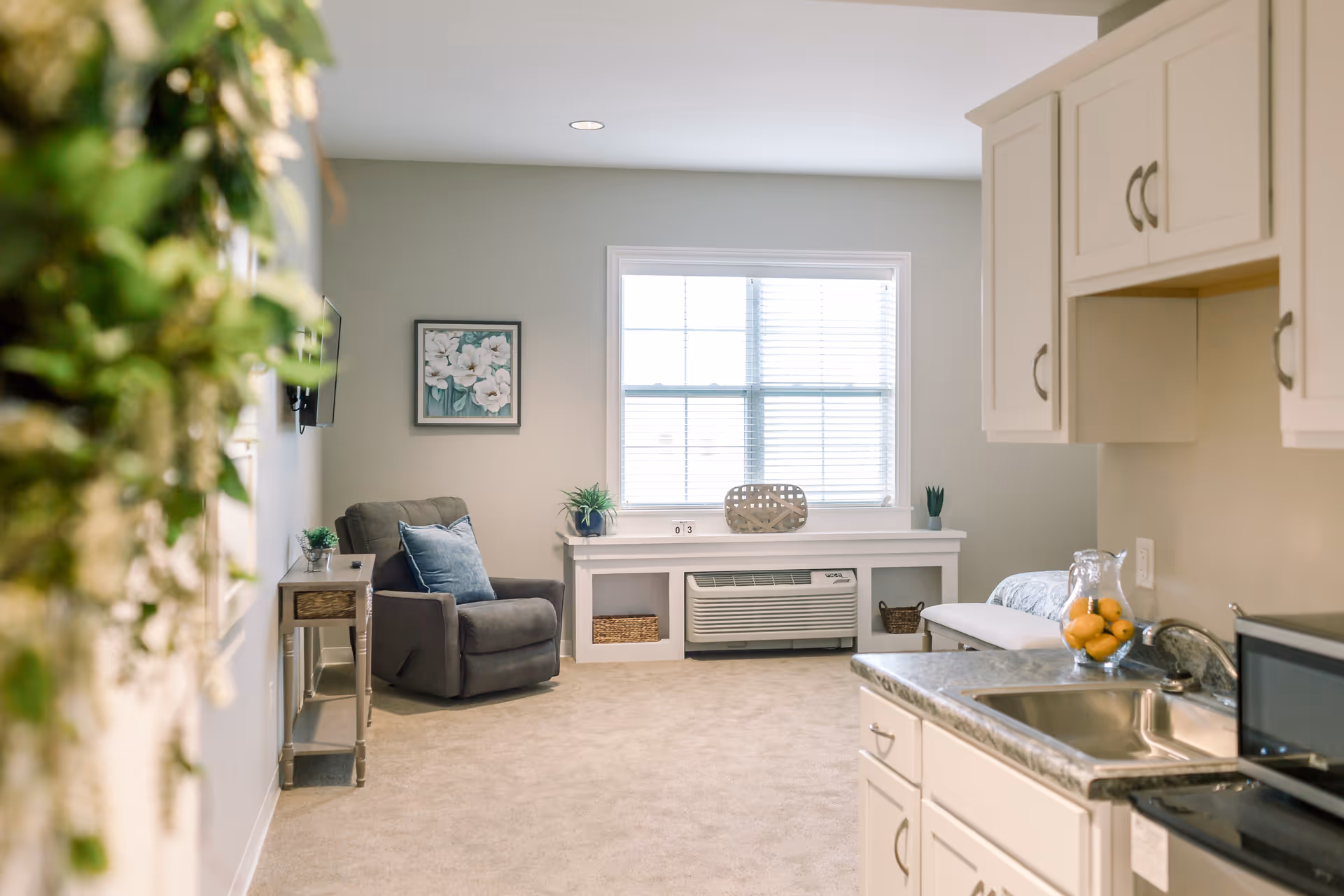 A cozy senior living room with a gray armchair, a small side table with a plant, a large window with blinds, and a built-in air conditioning unit below the window. The room has light-colored walls and carpet, with a kitchen area visible on the right side featuring a sink, countertop, microwave, and cabinets. There is also a decorative plant hanging on the left side of the image.