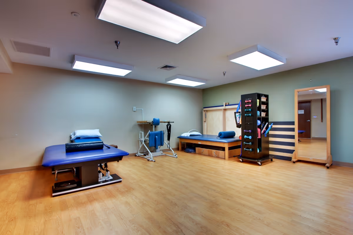 A bright therapy room with two treatment tables, exercise equipment, a rack of dumbbells and a full-length mirror.