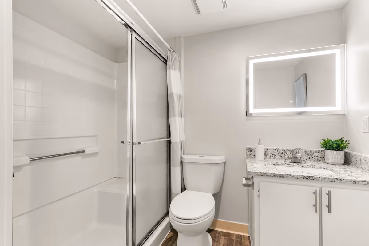 A modern bathroom featuring a glass shower, a toilet, and a granite countertop with a sink. The space is well-lit with a backlit mirror and has a neutral color scheme.