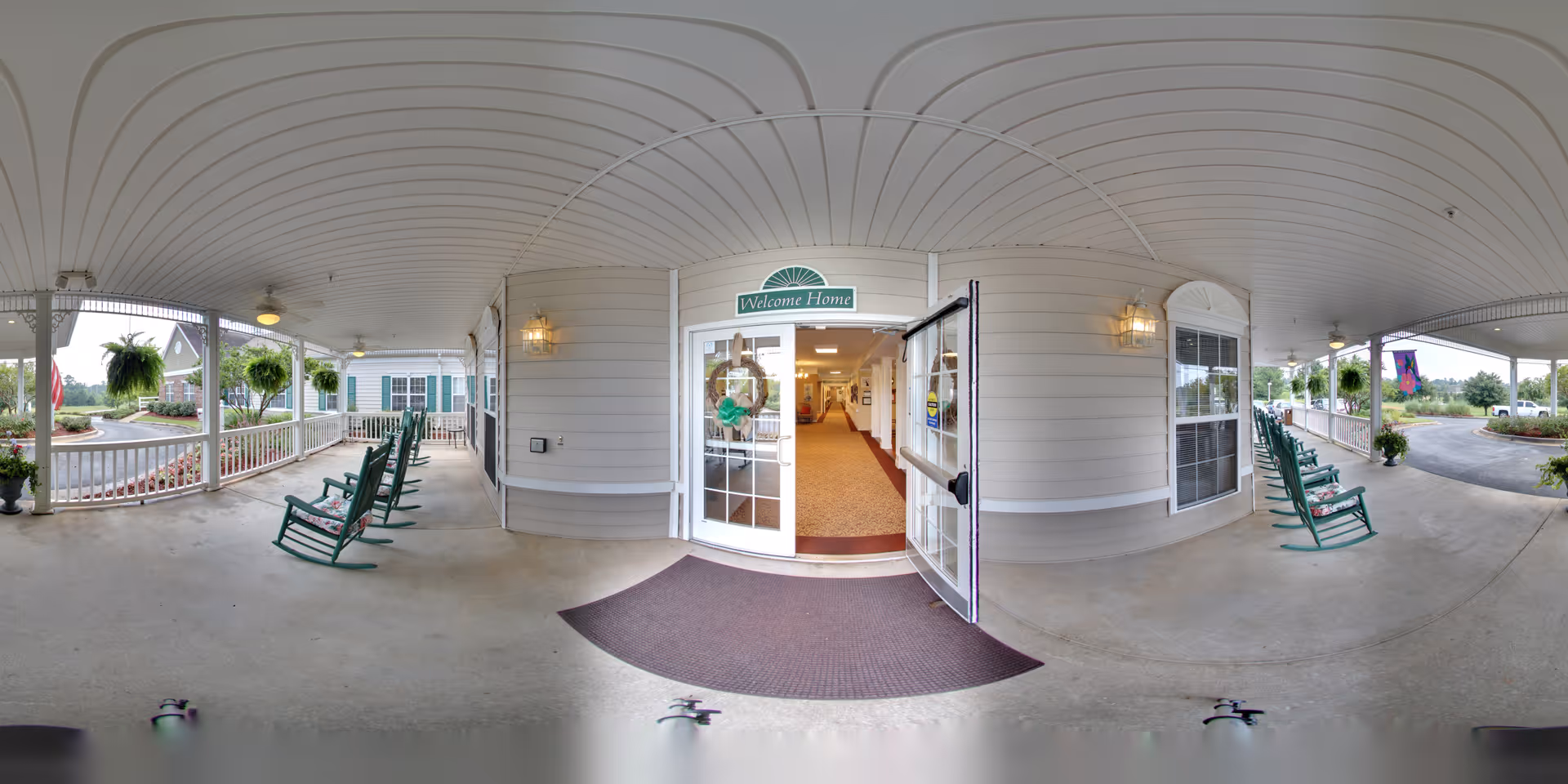 Covered front porch entrance with green rocking chairs, hanging ferns, and glass double doors under a 'Welcome Home' sign.