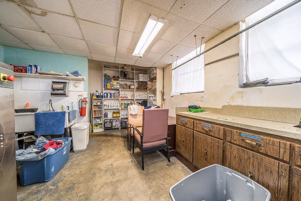 A utility or laundry room with a large sink, shelves stocked with various supplies, a table with chairs, and cabinets along one wall. There are two windows with white curtains, a blue plastic bin filled with clothes, and a trash bin. The floor appears worn and the walls show signs of wear.