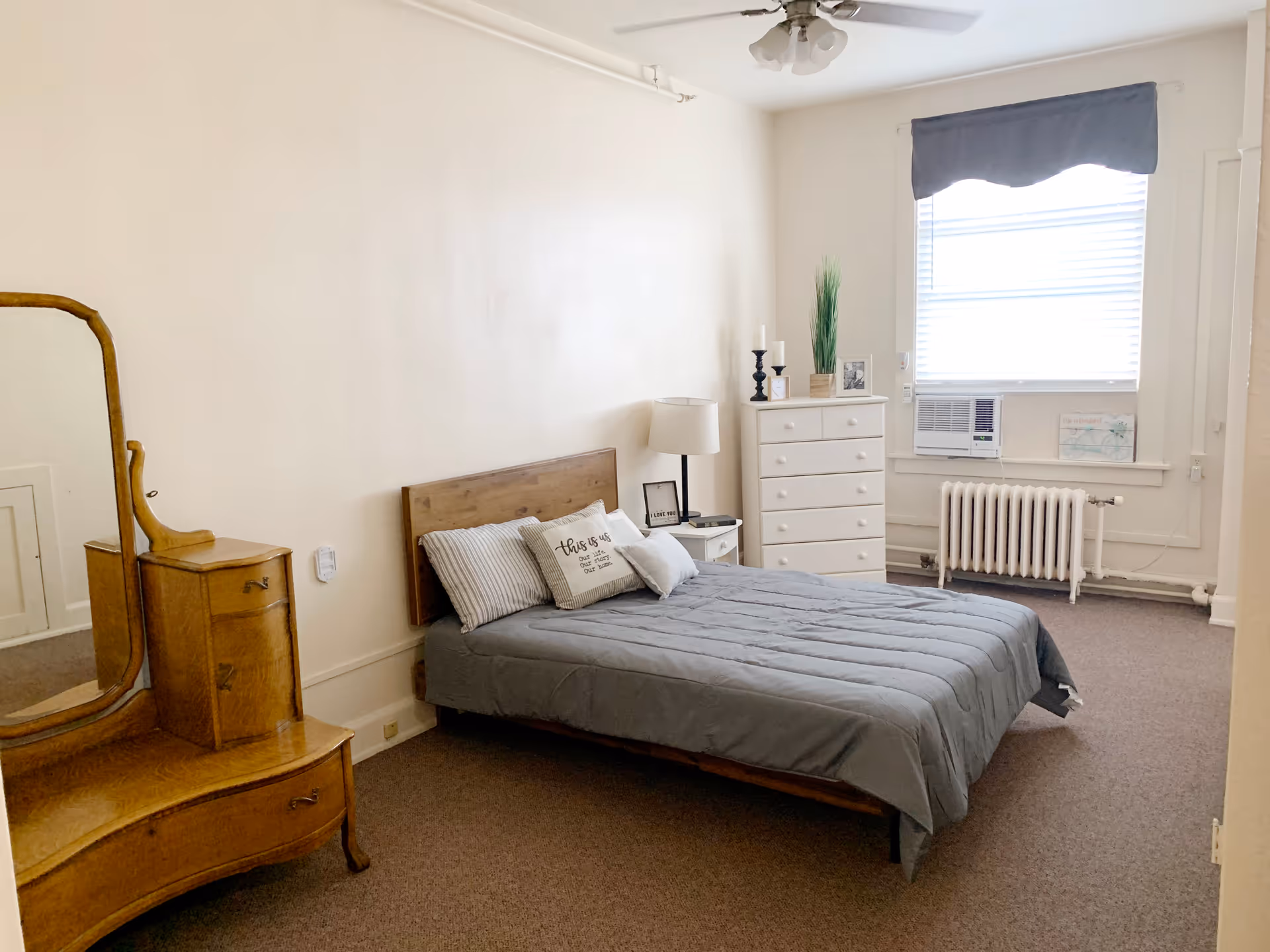 A simple bedroom with a wooden bed frame and gray bedding, three pillows including one with text, a white nightstand with a lamp and framed picture, a white chest of drawers with decorative items, a window with blinds and a blue valance, an air conditioning unit, a radiator, and a wooden vanity mirror with drawers.
