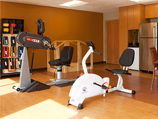 Interior exercise room with two recumbent bikes, an equipment rack, and kitchenette cabinets with a refrigerator in the background.