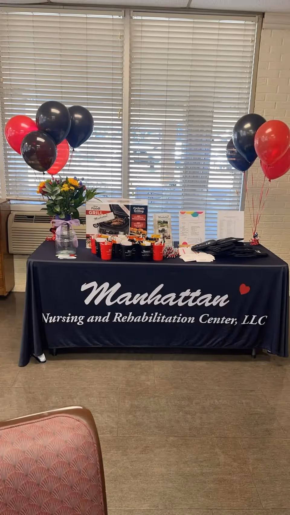 A decorated table inside a room with a dark blue tablecloth that reads 'Manhattan Nursing and Rehabilitation Center, LLC'. The table has red and black balloons on both sides, a vase with flowers, promotional materials, and some cups and papers arranged on top. Behind the table are large windows with closed blinds.