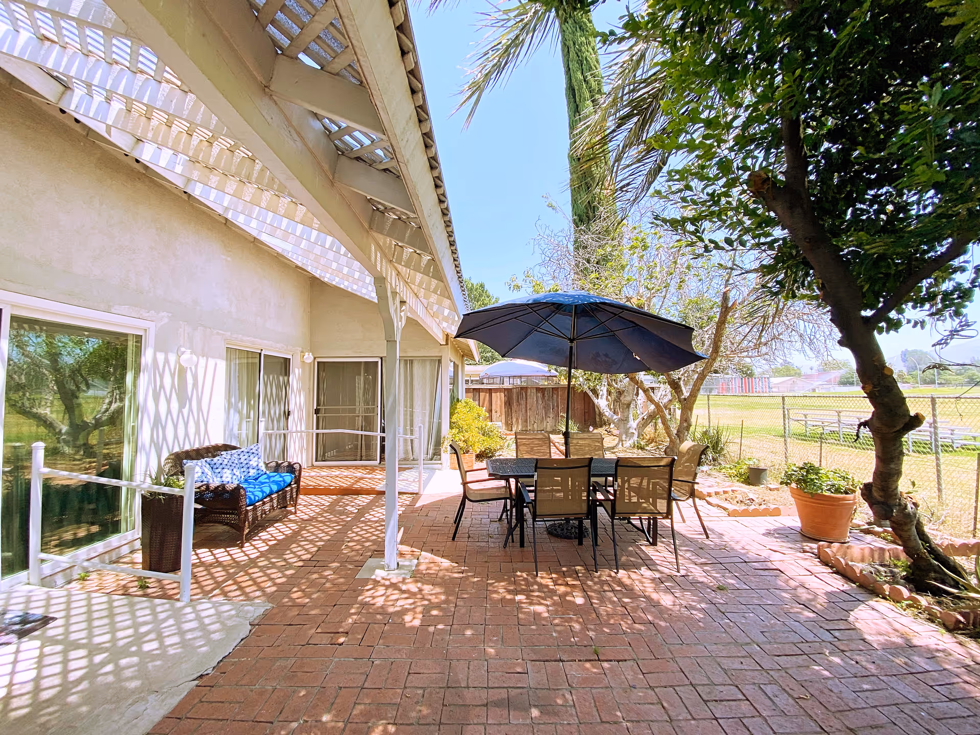 Outdoor patio area with brick flooring, a table with six chairs, and a large blue umbrella providing shade. There is a wicker bench with blue cushions against the building wall, which has sliding glass doors. Trees and plants surround the patio, and a chain-link fence encloses a grassy field in the background.
