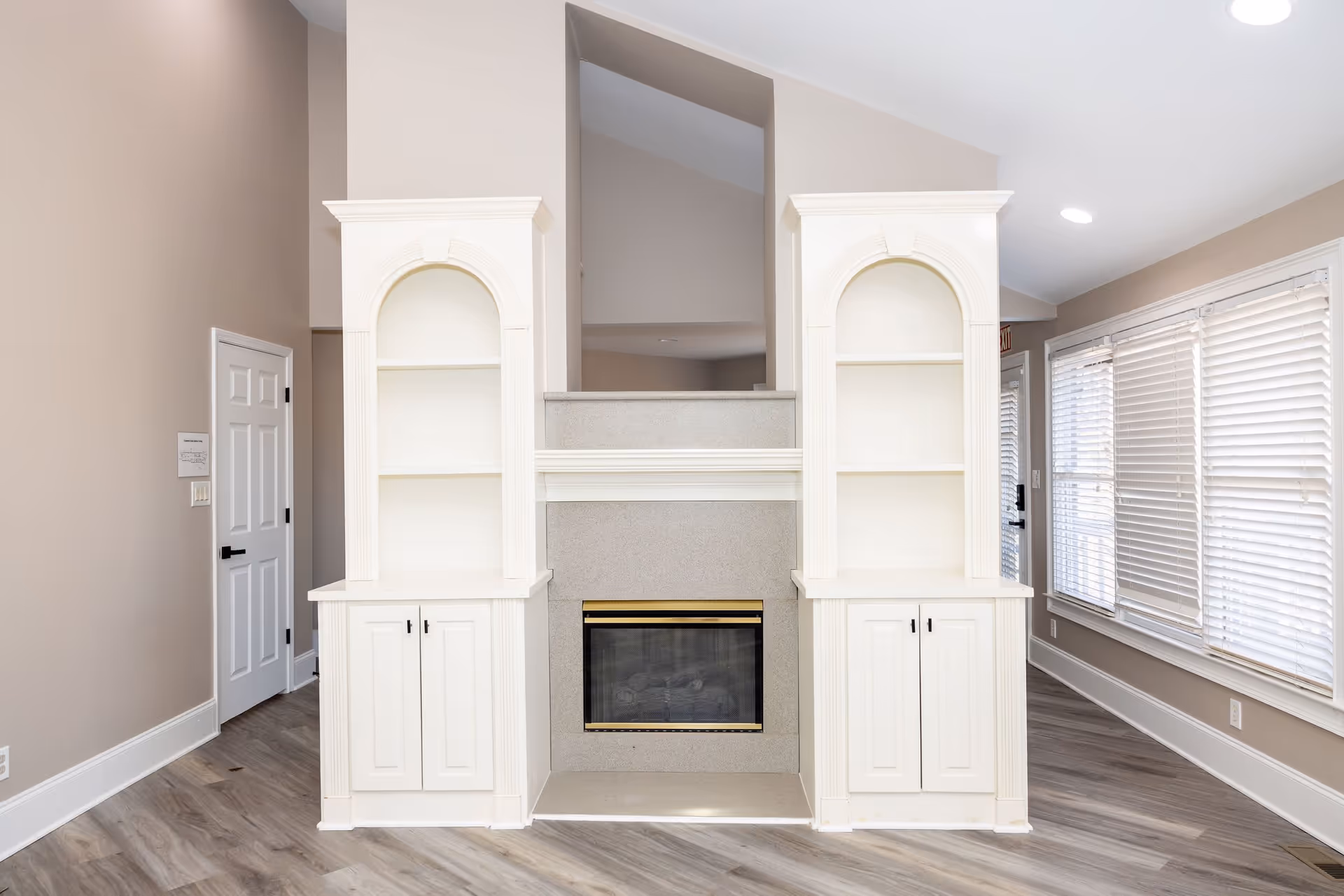 Interior view of a senior living facility room featuring a central fireplace with a gray stone surround and brass trim. On either side of the fireplace are white built-in shelves with arched tops and cabinets below. The room has light beige walls, wood flooring, and large windows with white blinds on the right side. A white door is visible on the left side of the image.