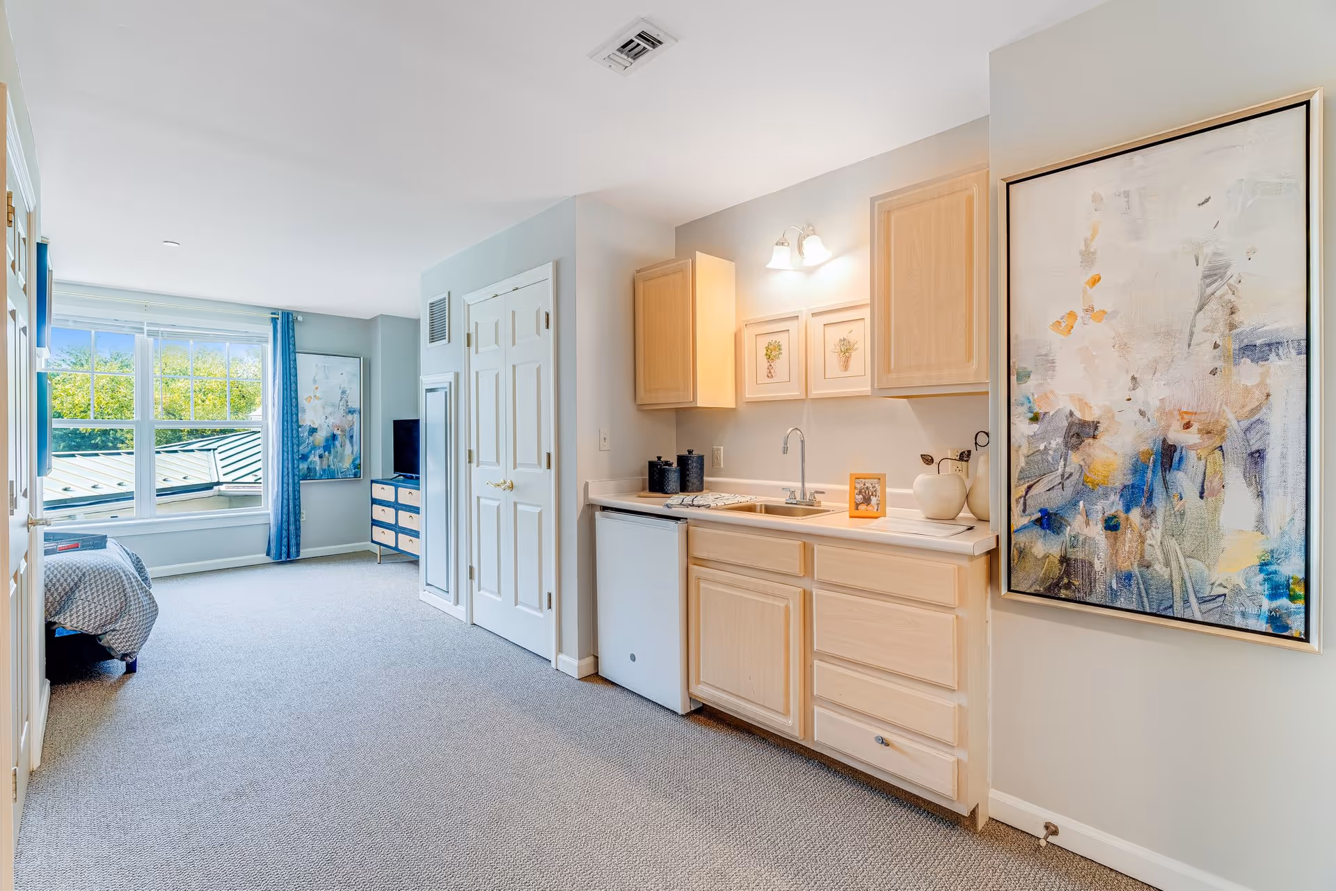 Interior view of a senior living facility room at Sunrise of Abington featuring a small kitchenette with light wood cabinets, a sink, and a mini refrigerator. The room has carpeted flooring and light-colored walls adorned with framed artwork. In the background, there is a bed near a large window with blue curtains, a dresser, and a TV. The space is bright with natural light coming through the window.