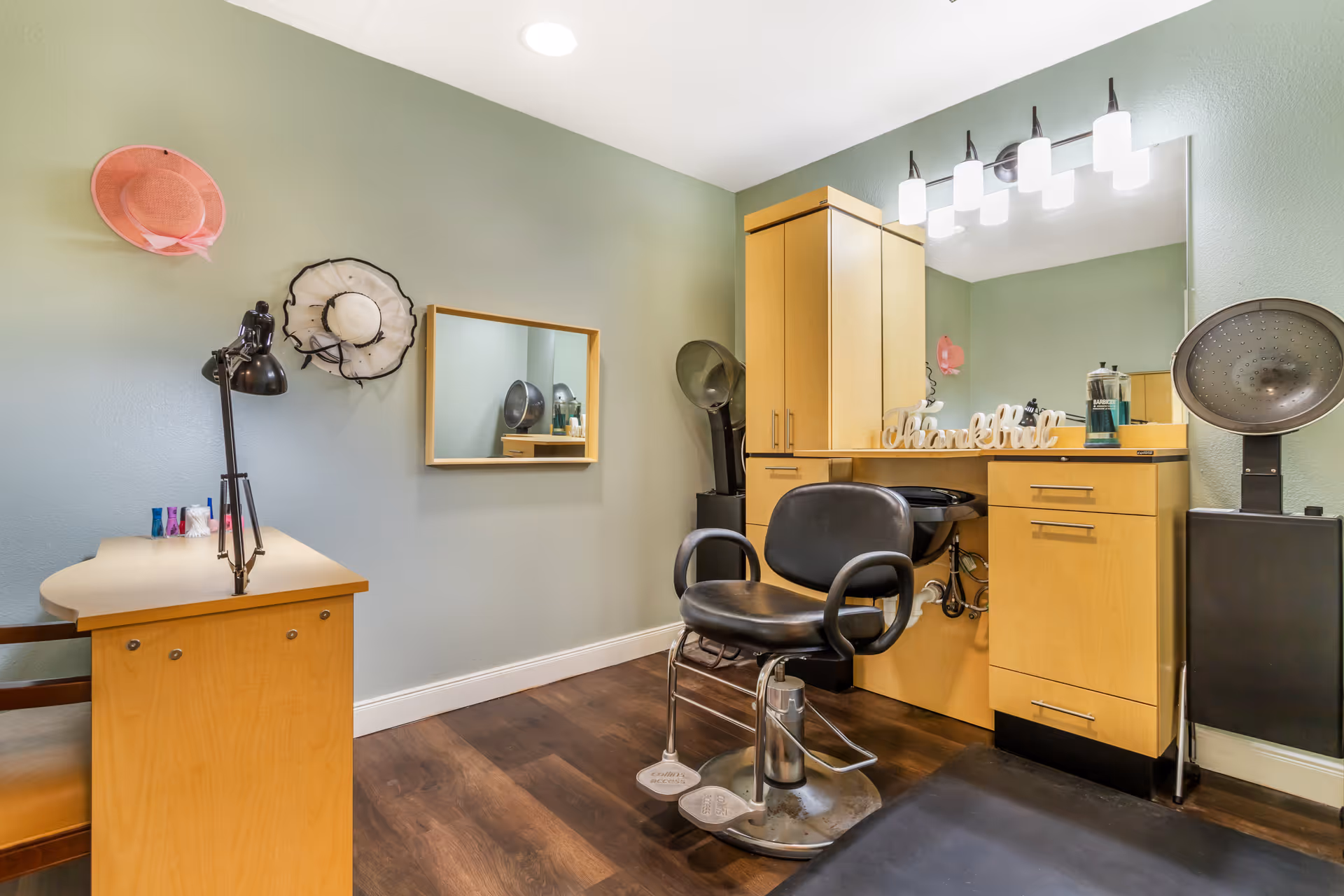 Hair salon station with a black styling chair, vanity mirror, wooden cabinets and hair dryers against a green wall.