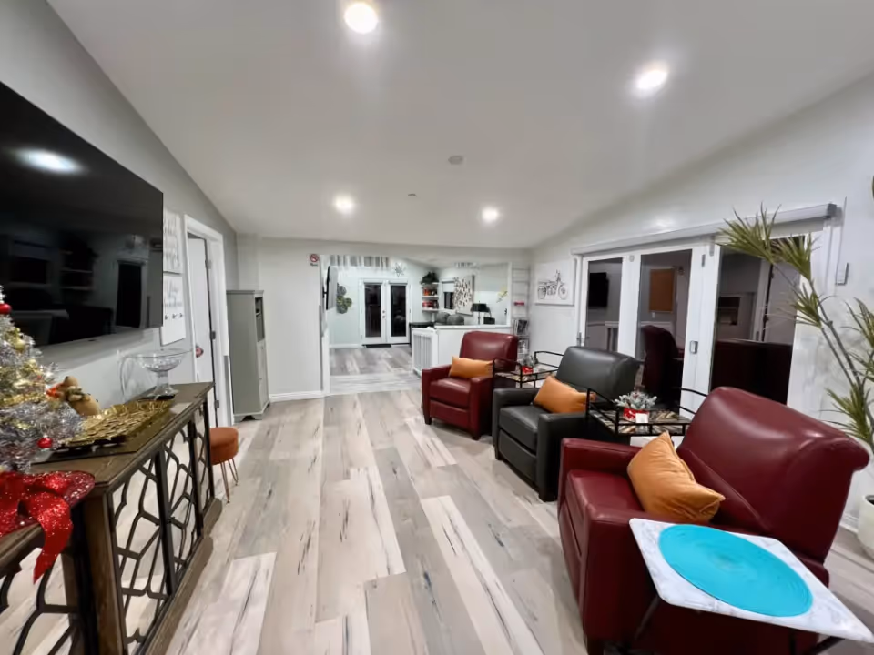 Bright communal living room with red and gray armchairs, a wall-mounted TV, side tables, and holiday decorations.