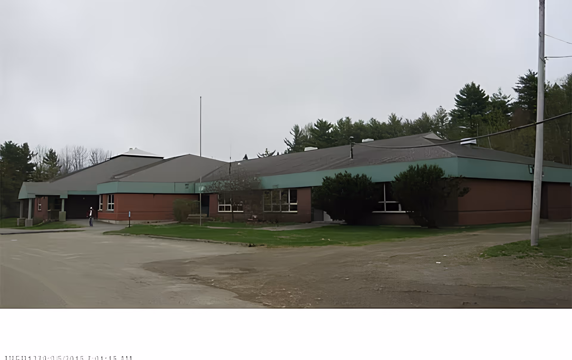Exterior view of a single-story brick building with a green roof trim, surrounded by grass and trees under an overcast sky. A person is standing near the entrance on the left side of the building.