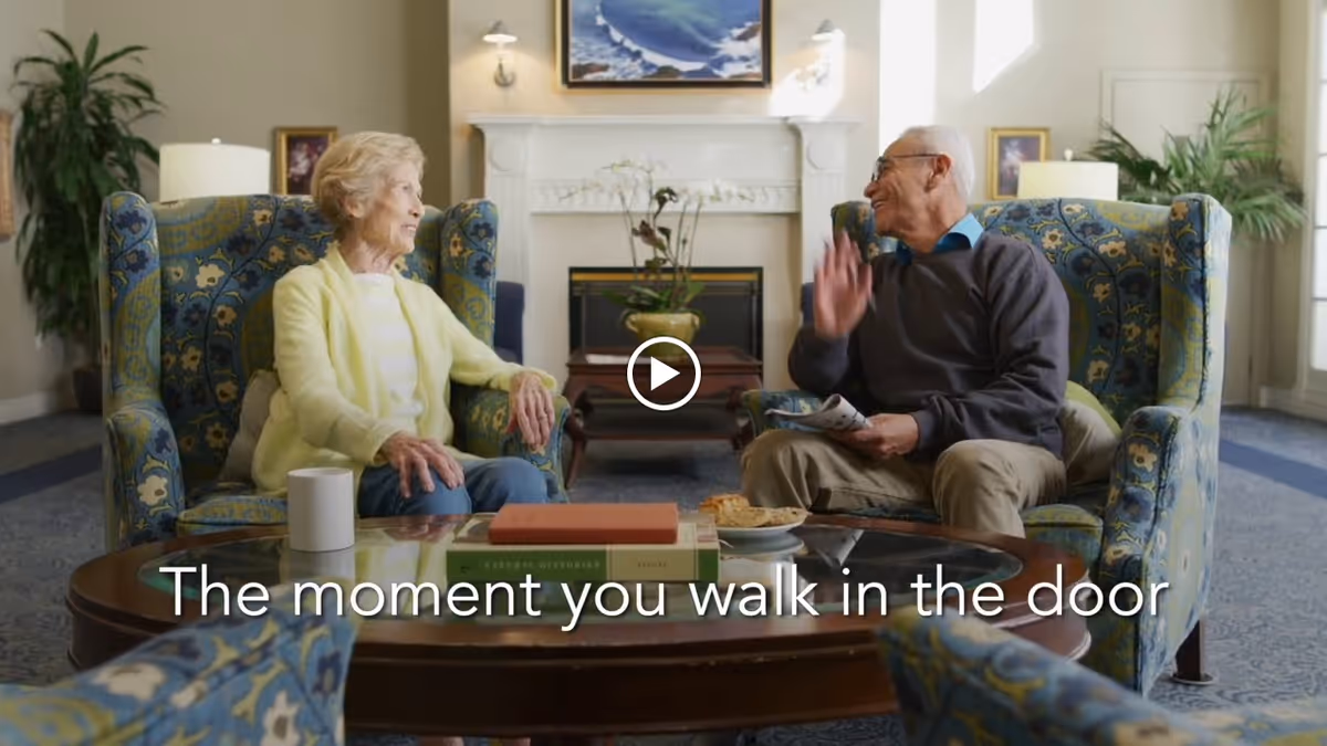Two elderly people sitting in patterned armchairs in a cozy living room with a fireplace, plants, and framed pictures on the walls, engaged in a friendly conversation. A coffee table with books, a cup, and a plate of cookies is in front of them. Text on the image reads 'The moment you walk in the door.'