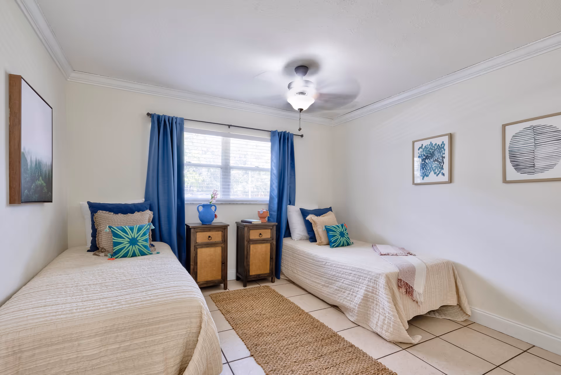 A bright bedroom with two single beds placed parallel to each other, each bed covered with a beige quilt and decorated with multiple pillows in blue, beige, and green colors. Between the beds are two wooden nightstands with a blue vase and a small orange pot on top. A window with blue curtains is centered on the back wall, and two framed abstract artworks hang on the walls. The floor is tiled with a woven beige rug running between the beds. A ceiling fan with a light is turned on above.