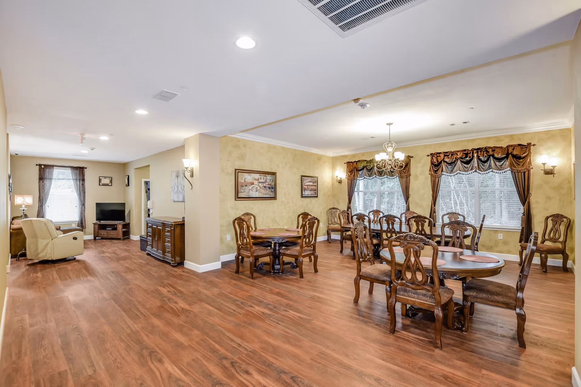 A spacious, well-lit interior room with wooden flooring featuring multiple round dining tables with ornate wooden chairs. The walls are painted in a warm beige tone with framed artwork and decorative wall sconces. Large windows with brown curtains allow natural light to enter. In the background, there is a small sitting area with a beige recliner, a brown sofa, a TV on a wooden stand, and a side table with a lamp.