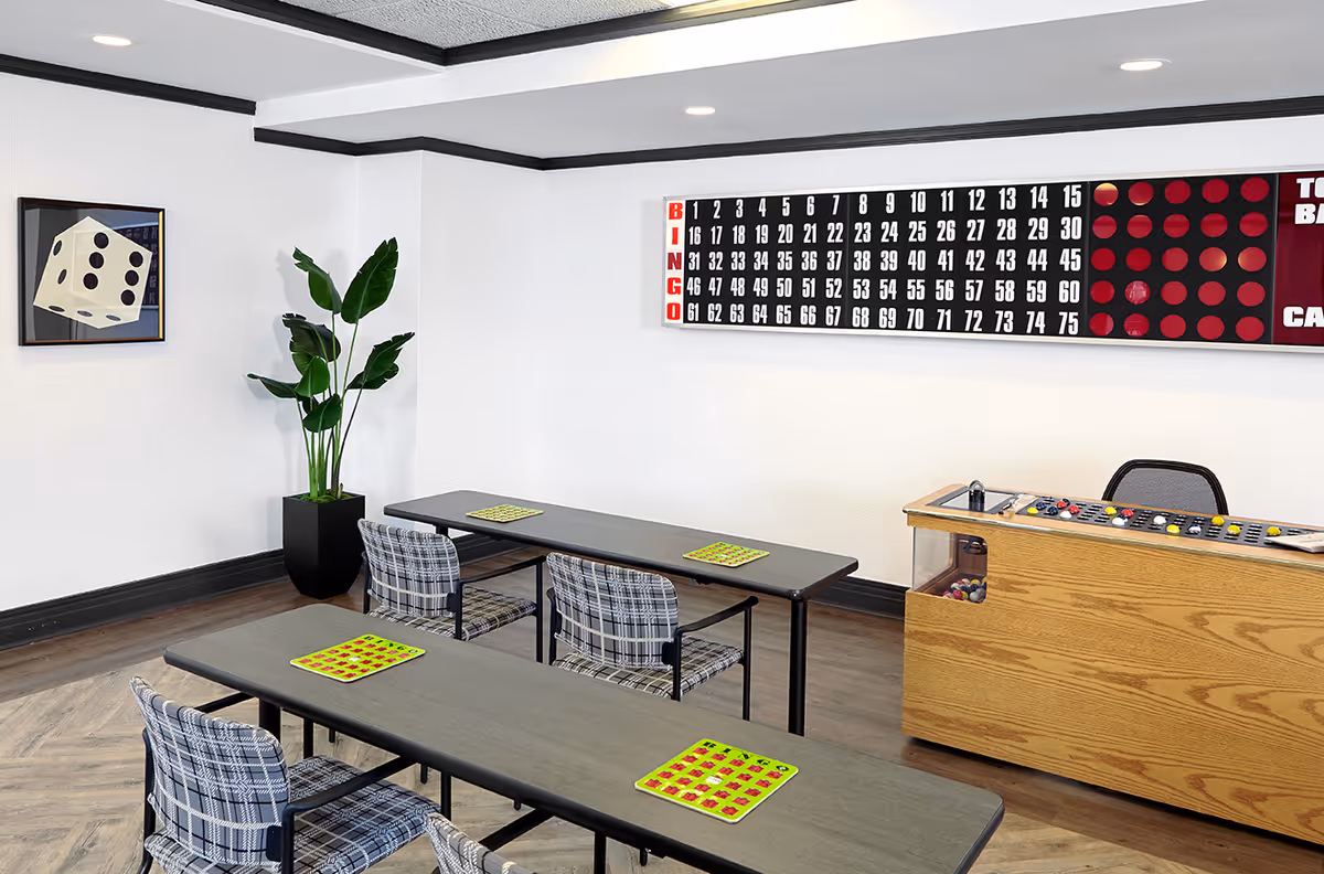 A game room with two tables and plaid chairs arranged for bingo. Each table has bingo cards placed on it. On the wall, there is a large bingo board and a wooden bingo machine with colorful balls inside. A framed picture of a large die and a tall green plant in a black pot are also visible.