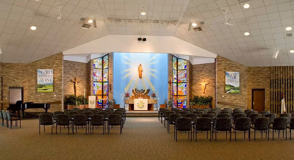 Interior view of a chapel with rows of chairs facing an altar flanked by stained glass windows and religious artwork.