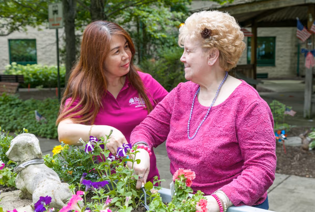 A caregiver and an elderly woman are gardening together outdoors. The caregiver, wearing a magenta shirt with the Elmwood Hills Healthcare Center logo, is assisting the elderly woman who is wearing a pink sweater and a beaded necklace. They are surrounded by colorful flowers and greenery, with a stone dog statue nearby.